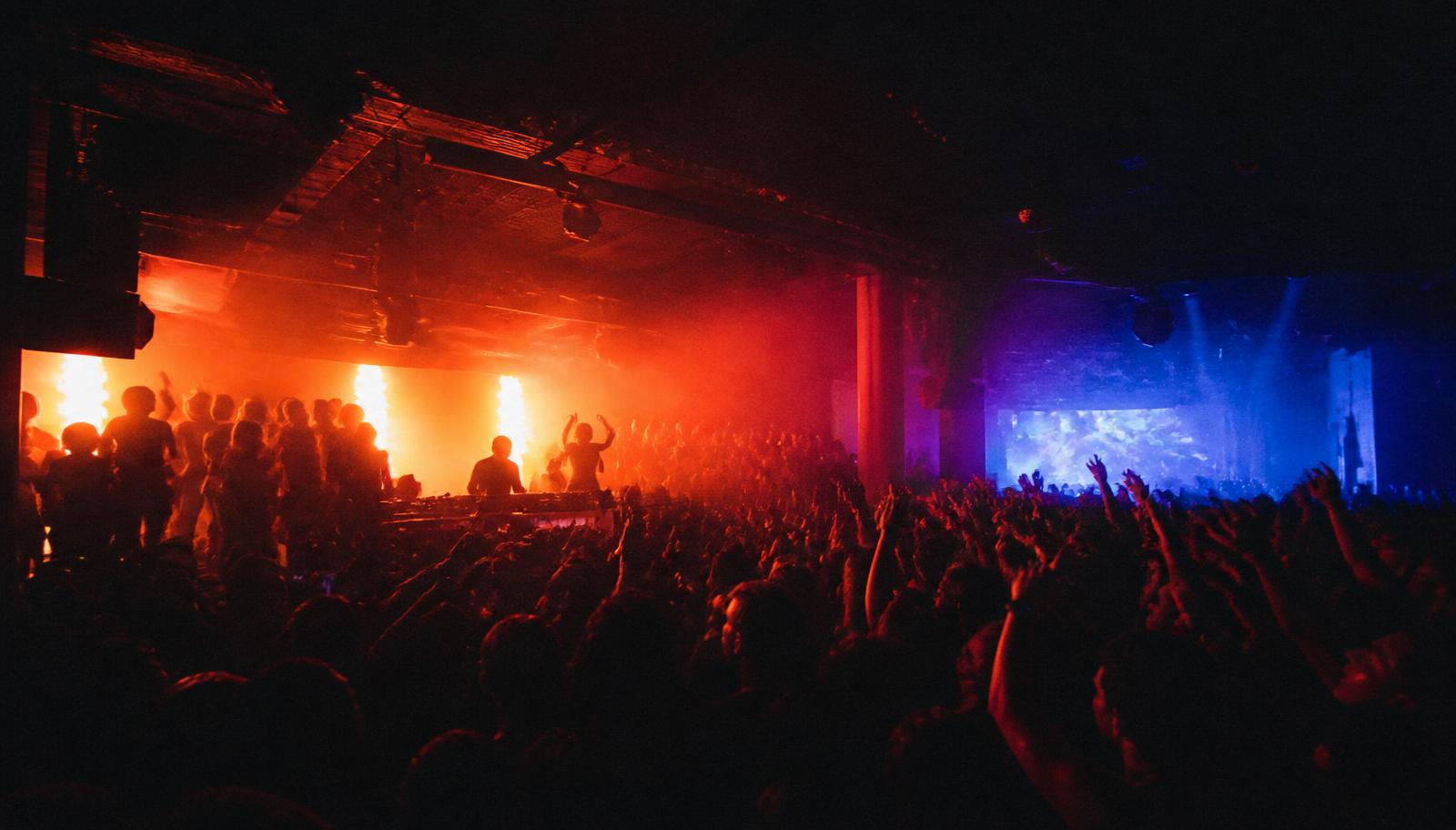 Smoke-filled club with two stages, red and blue lights, crowd in perreo